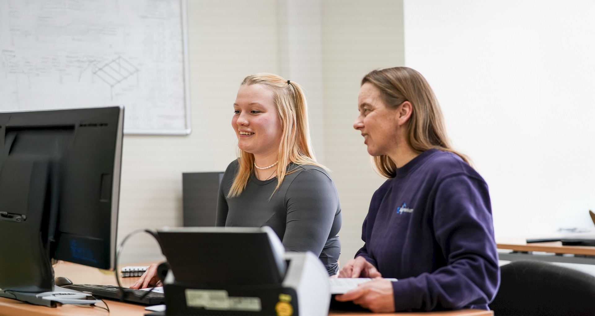 Schülerin und Mitarbeiterin sitzen gemeinsam am Computer im Büro von TS Aluminium und arbeiten an einem Projekt.