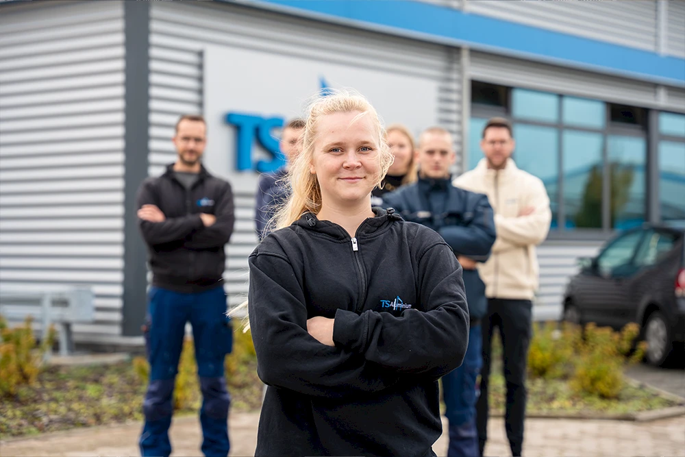 Auszubildende steht selbstbewusst mit verschränkten Armen vor dem Team vor dem Firmengebäude von TS Aluminium.