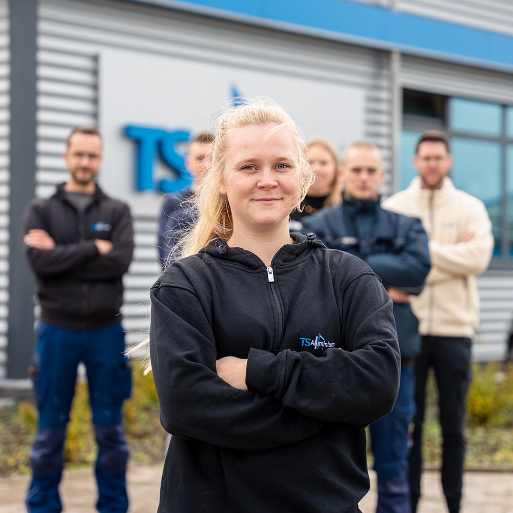 Junge Frau im TS Aluminium-Hoodie steht selbstbewusst mit verschränkten Armen vor einer Gruppe junger Kolleginnen und Kollegen vor dem Firmengebäude von TS Aluminium.