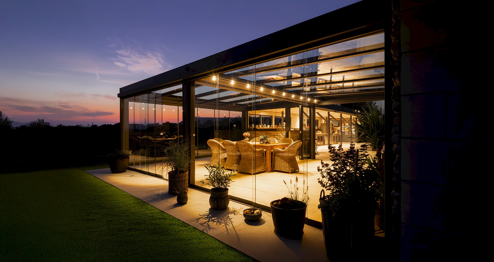 Moderner Sommergarten mit Glaswänden und stimmungsvoller LED-Beleuchtung bei Sonnenuntergang, eingerichtet mit gemütlichen Rattanmöbeln.