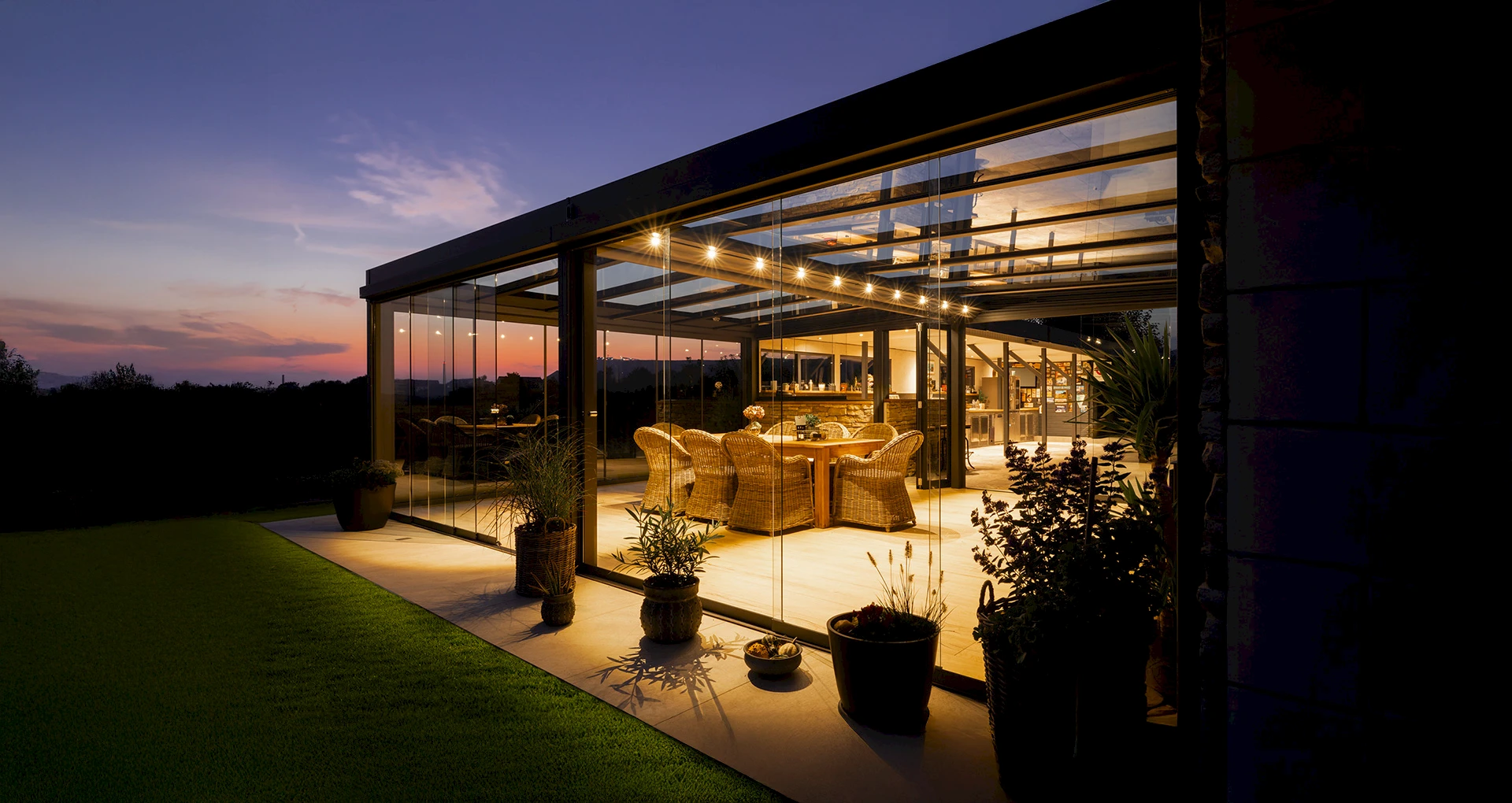 Moderner Sommergarten mit Glaswänden und stimmungsvoller LED-Beleuchtung bei Sonnenuntergang, eingerichtet mit gemütlichen Rattanmöbeln.