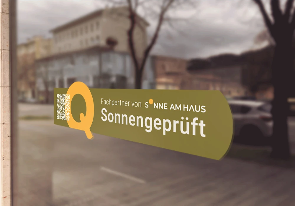Aufkleber mit Logo und Schriftzug „Fachpartner von Sonne am Haus – Sonnengeprüft“ auf einer Fensterscheibe, im Hintergrund unscharfe Straße mit Gebäuden und parkendem Auto