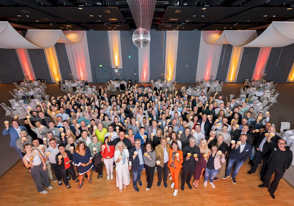 Große Gruppe von Menschen in einem festlich dekorierten Saal, die runde Sonne am Haus Bierdeckel in die Kamera halten